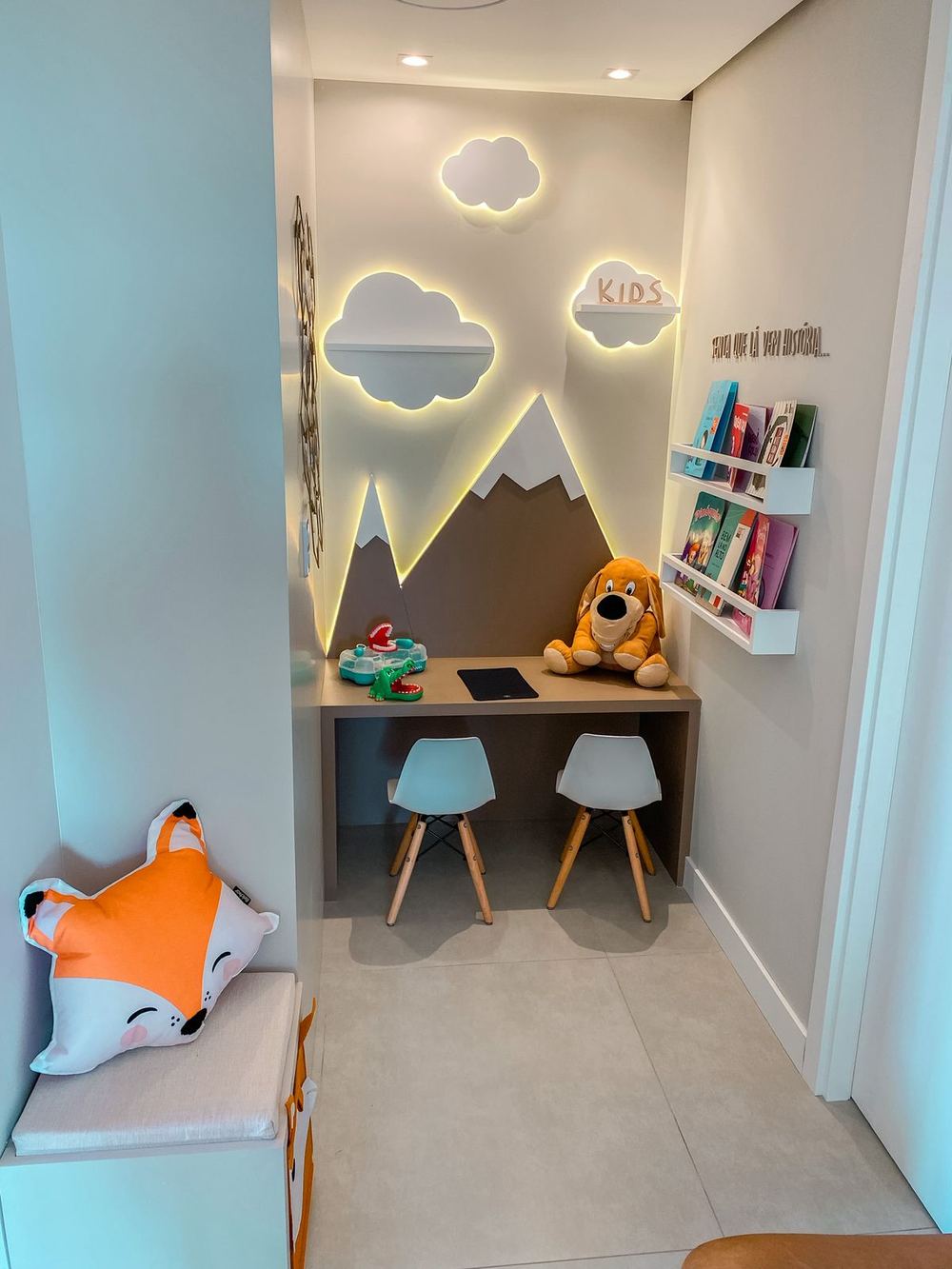 Área kids com nuvens iluminadas, montanhas na parede, mesinha, cadeiras infantis e livros