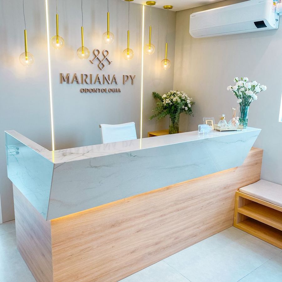 Recepção da clínica com balcão em mármore e madeira, luminárias douradas e o logotipo Mariana Py Odontologia na parede