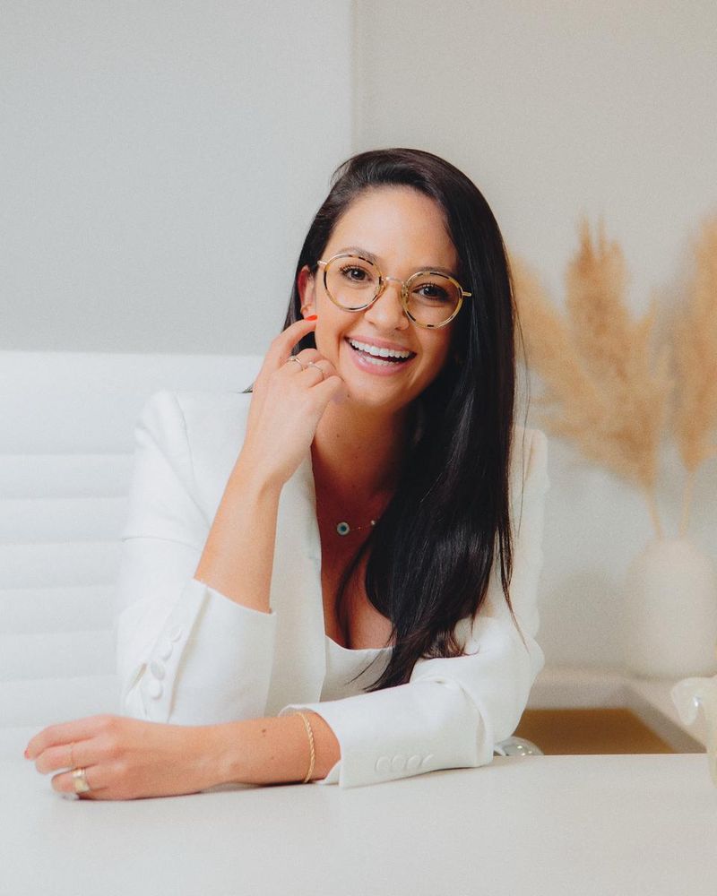 Dra. Mariana Py, sorridente, de óculos e blazer branco, sentada à mesa do consultório