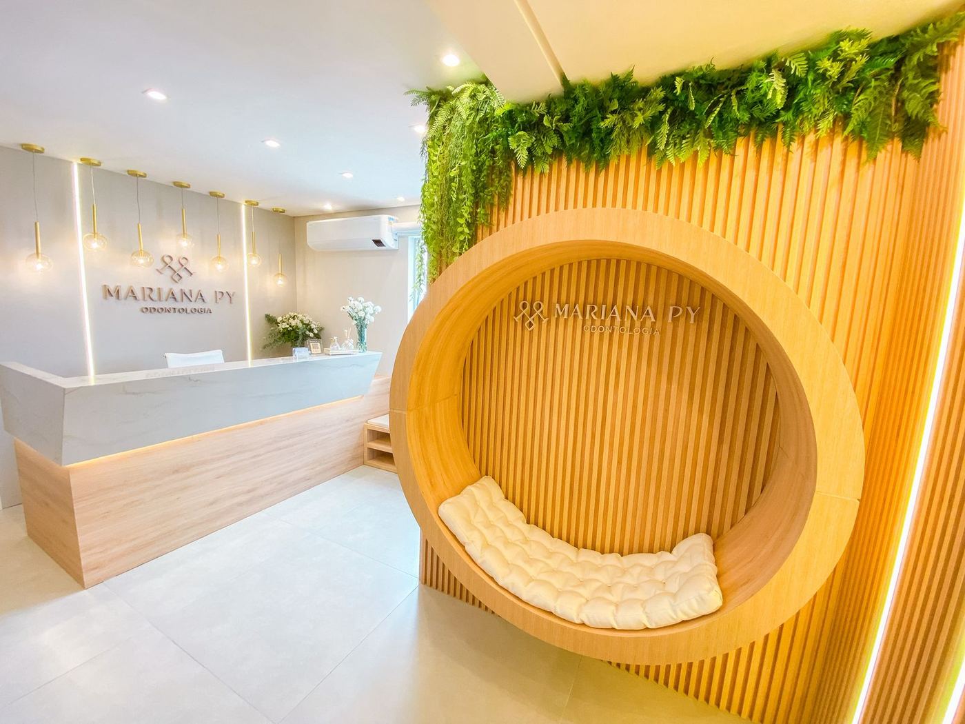 Recepção da clínica Mariana Py Odontologia, com balcão claro, luminárias douradas e um banco circular de madeira com almofada branca