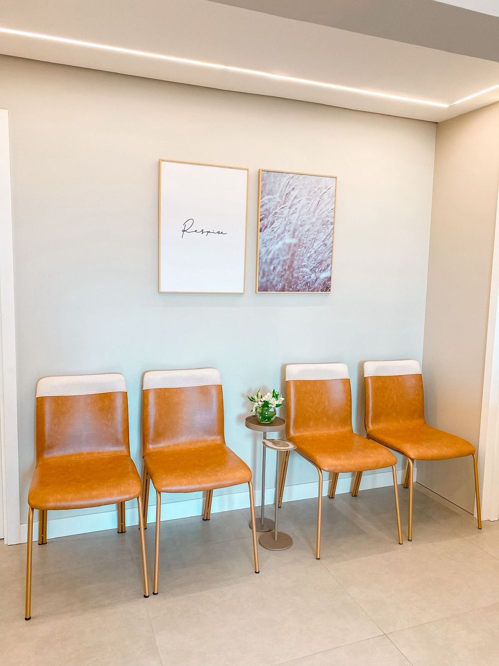 Sala de espera com cadeiras caramelo, mesinha com flores e quadros na parede